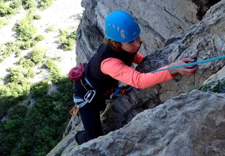Une grimpeuse en casque bleu progresse sur une paroi rocheuse, corde bleue en main. L'escalade à Thaurac offre des vues spectaculaires.