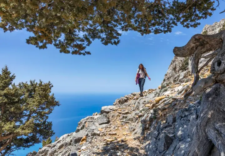 Une randonneuse explore un sentier rocailleux sur une montagne de Crète, avec la mer bleue en arrière-plan.