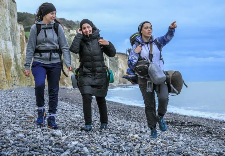 Trois randonneuses marchent sur une plage de galets, le ciel est couvert. L'une d'elles pointe le doigt vers l'horizon, peut-être pour indiquer la fin de leur GR 21 étapes.