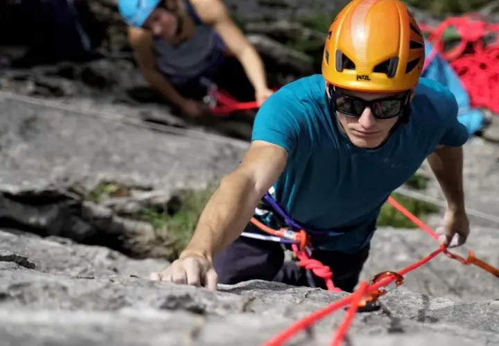 Un grimpeur concentré, casque orange, s'engage dans une voie d'escalade 64. Une corde rouge vif l'assure.