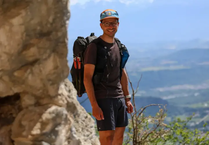 Jérémy Bigé, souriant, prêt pour l'aventure avec son sac à dos et ses bâtons de randonnée, dominant un paysage de montagnes.