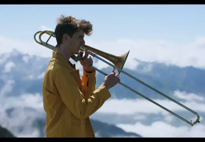 Victor Decamp Destivelle, un jeune homme aux cheveux ébouriffés, joue du trombone dans un paysage montagneux brumeux.
