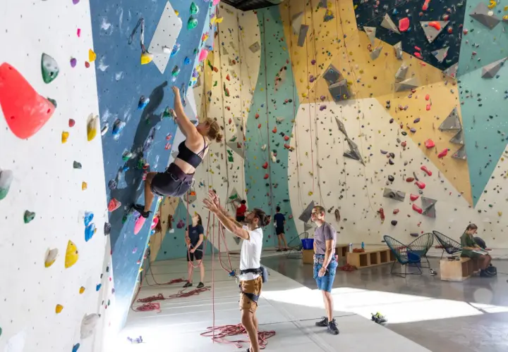 Une femme fait de l'escalade climb up sur un mur coloré, d'autres personnes observent ou se préparent.