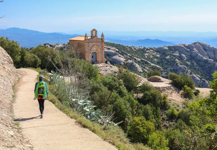 Une randonnée à Montserrat, avec une femme portant un sac à dos et une corde, menant à une petite chapelle perchée sur une colline rocheuse.