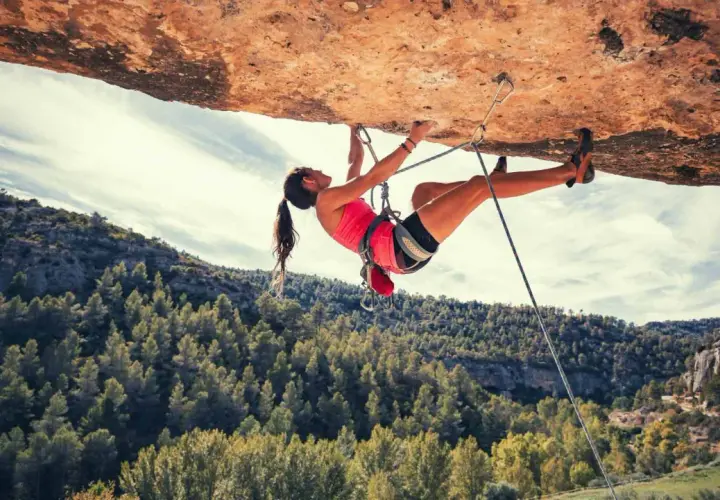 Une femme en pleine escalade extrême, suspendue sous un surplomb rocheux, avec une forêt verdoyante en arrière-plan.