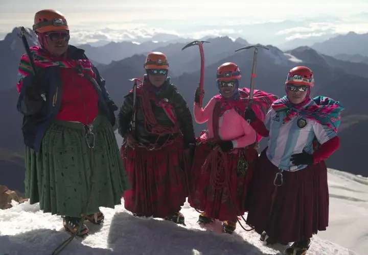 Quatre cholitas escaladoras, casques et piolets, posent fièrement sur un sommet enneigé, leurs jupes colorées flottant au vent.