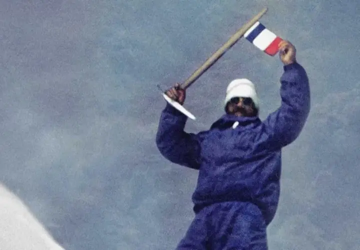 Les mains de Maurice Herzog brandissent un drapeau français au sommet d'une montagne enneigée, symbole d'une victoire alpine.