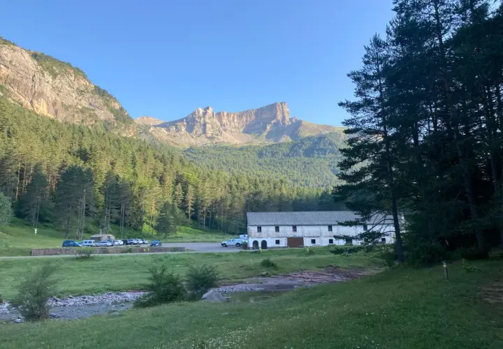 Vue panoramique de la **selva de Oza** avec une bâtisse blanche, des montagnes imposantes et une forêt dense sous un ciel bleu clair.
