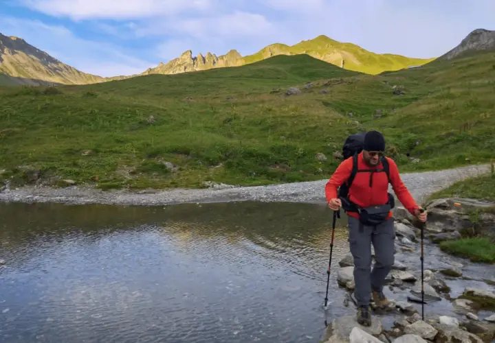 Un randonneur traverse un ruisseau en utilisant des bâtons de marche. Le soleil éclaire les montagnes verdoyantes en arrière-plan.