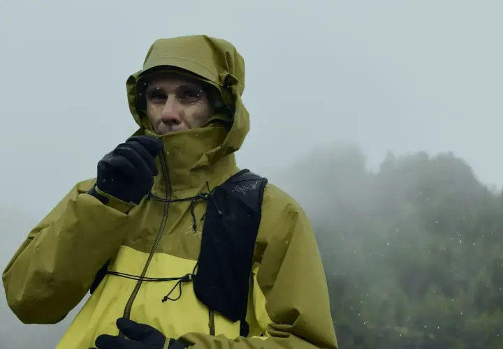 Un homme en veste Gore-Tex jaune et verte, sous la pluie, ajuste son col. Le lavage de cette veste est essentiel pour maintenir son imperméabilité.