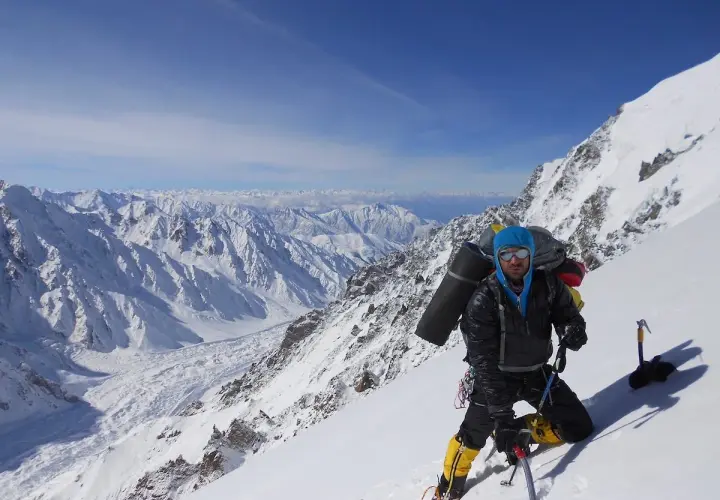 Daniele Nardi, alpiniste, en pleine ascension sur une pente enneigée, avec un sac à dos imposant et des crampons. Le paysage montagneux est spectaculaire.