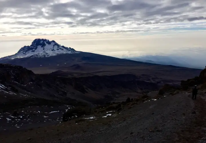 Vue sommet Kilimandjaro, paysage désertique et enneigé sous un ciel nuageux. Un randonneur solitaire sur le sentier.