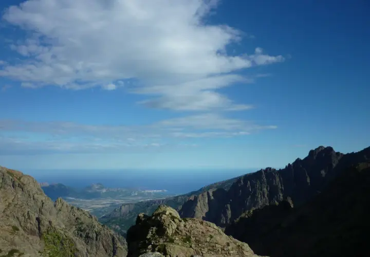 Vue spectaculaire sur la mer depuis les montagnes escarpées du GR20 partie nord. Le ciel bleu est parsemé de nuages blancs.