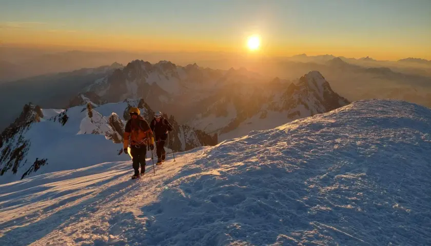Deux alpinistes progressent sur un sentier enneigé au lever du soleil, dominant un paysage de montagnes spectaculaire. Le sommet montagneux est baigné d'une lumière dorée.