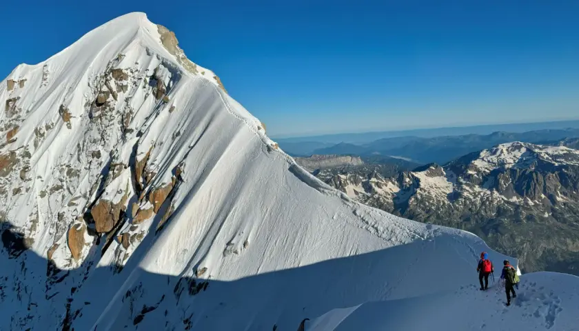 Deux alpinistes escaladent la crête enneigée de l'Aiguille Verte, sous un ciel bleu intense. Le soleil projette de longues ombres sur la montagne.