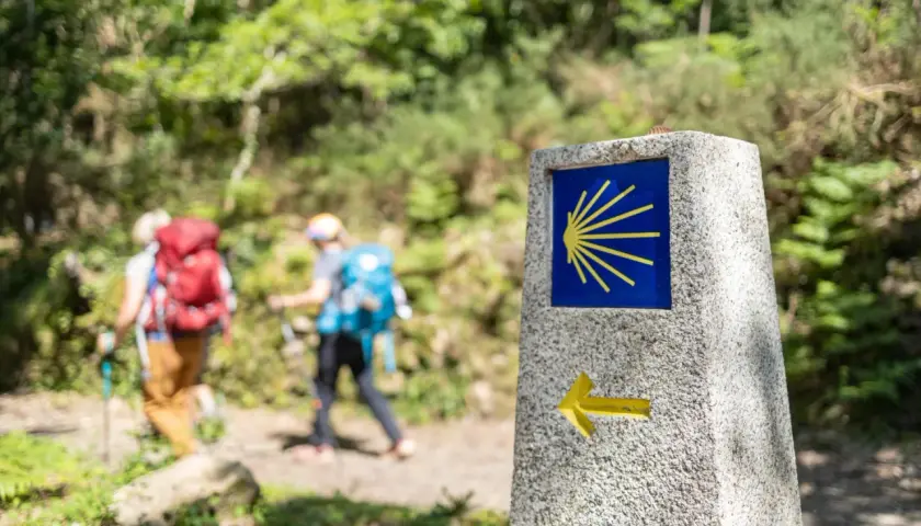 Panneau du Camino de Santiago avec une coquille jaune sur fond bleu et une flèche. Deux randonneurs avancent sur le chemin.