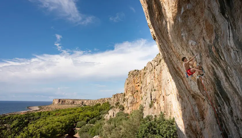 Un grimpeur défie la falaise en Sicile, avec la mer et la végétation en toile de fond. L'escalade sicile offre des vues spectaculaires.