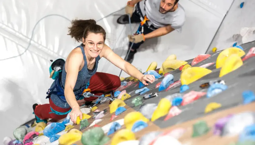 Une femme souriante en tenue d'escalade grimpe un mur coloré, assurée par un homme en arrière-plan.