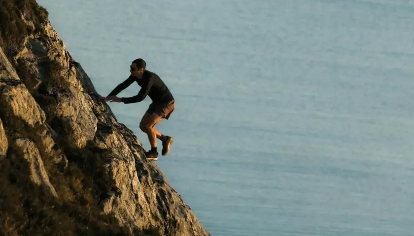 Kilian Jornet escalade une paroi rocheuse abrupte, dominant un plan d'eau calme, rappelant l'esprit d'aventure dans les Pyrénées.