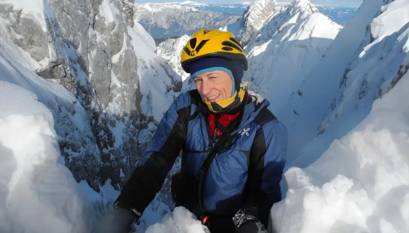 Une alpiniste souriante, Nives Meroi, en plein effort sur une crête enneigée, casque jaune, montagnes majestueuses en arrière-plan.