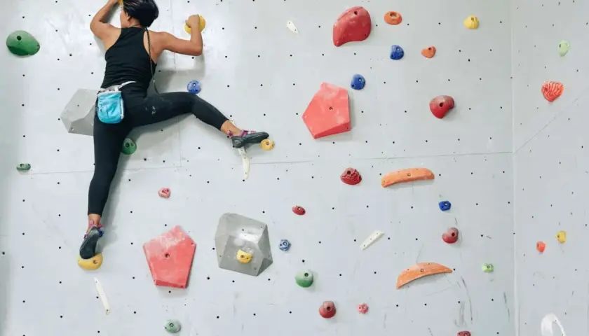 Grimpeuse en action, un pied dans la main, sur un mur d'escalade coloré. Elle progresse avec agilité.