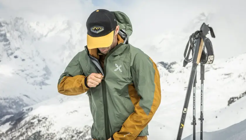 Un skieur ajuste sa veste technique, peut-être pour comparer le confort entre Primaloft vs Gore-Tex, avec des montagnes enneigées en arrière-plan.