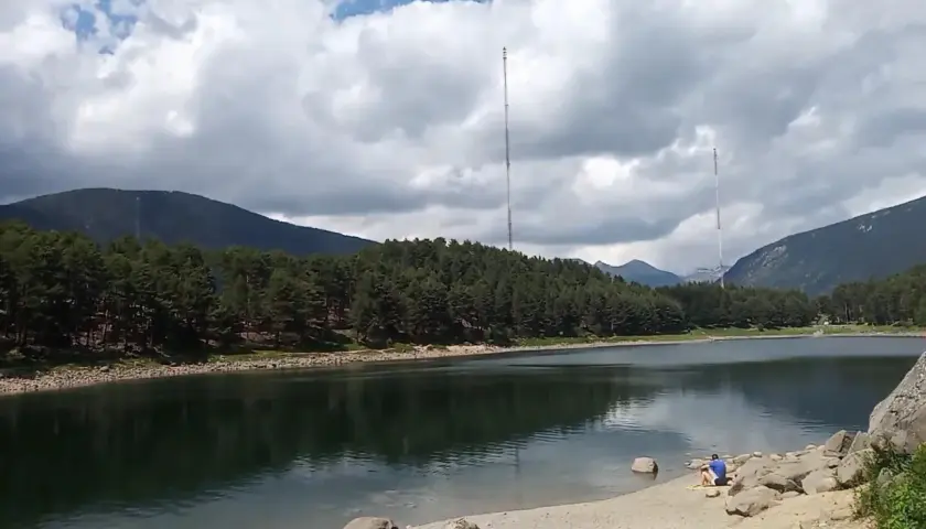 Vue paisible du lac d'Engolasters, entouré de forêts de pins et de montagnes. Deux tours de transmission se dressent à l'horizon.