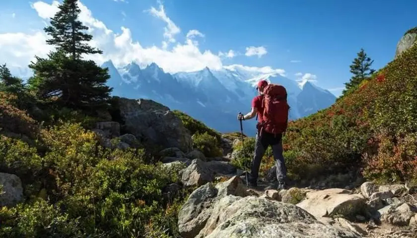 Un randonneur solo, sac à dos rouge, bâtons de marche, admire les montagnes majestueuses sous un ciel bleu éclatant.