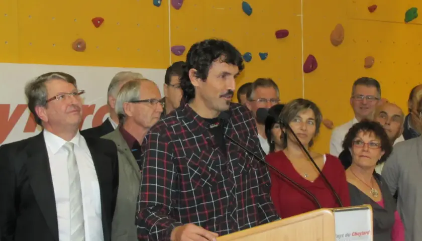 Antoine Cayrol, orateur charismatique, s'exprime devant un mur d'escalade coloré, entouré d'une foule attentive.