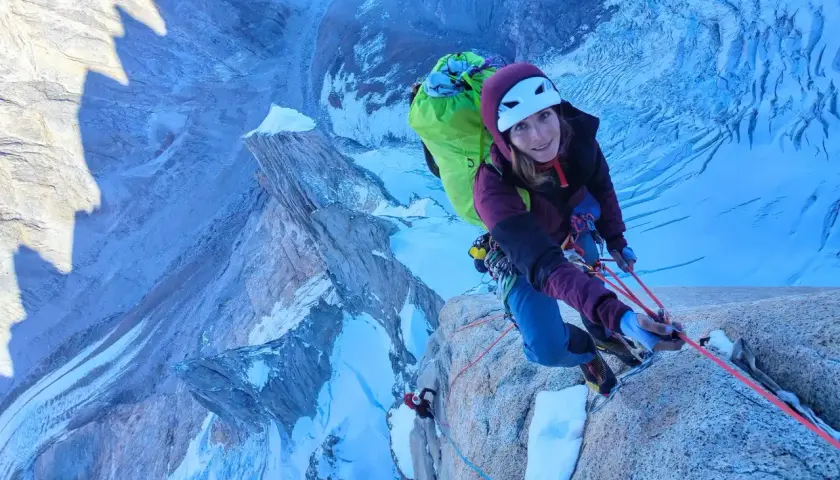 Lise Billon, casque blanc, grimpeuse souriante, est en pleine ascension sur une paroi rocheuse glacée, avec un sac à dos vert vif.