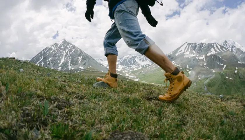 Un randonneur en bottes jaunes gravit une pente herbeuse, les montagnes enneigées en toile de fond. Une belle rando montagne.