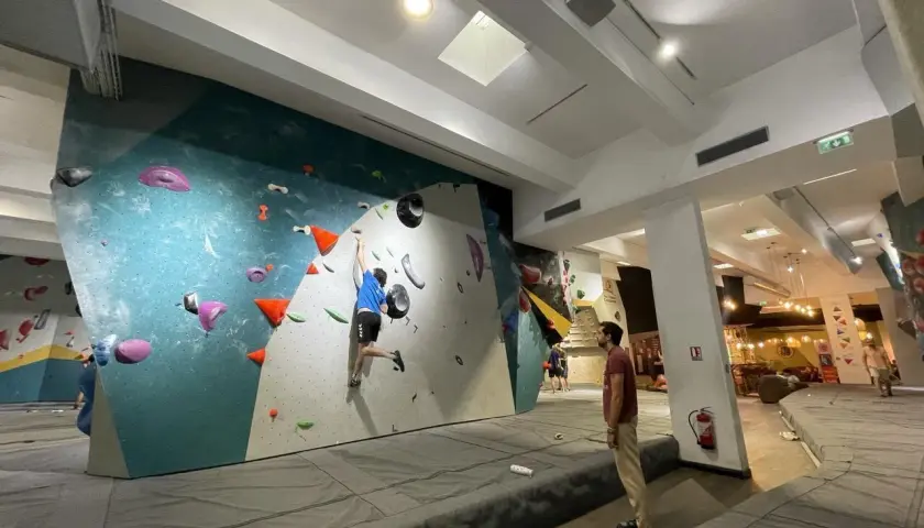 Un jeune homme en bleu pratique l'escalade sur un mur coloré, un autre spectateur l'observe. L'ambiance est propice à l'escalade nation.
