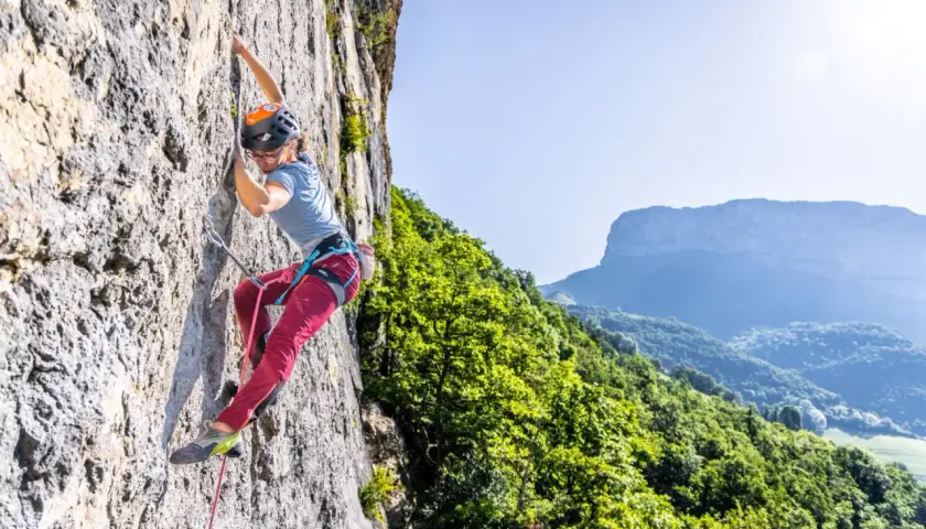 Une grimpeuse évolue sur une paroi rocheuse, profitant d'une journée ensoleillée dans ce parc d'escalade naturel. Le paysage montagneux verdoyant offre un panorama spectaculaire.
