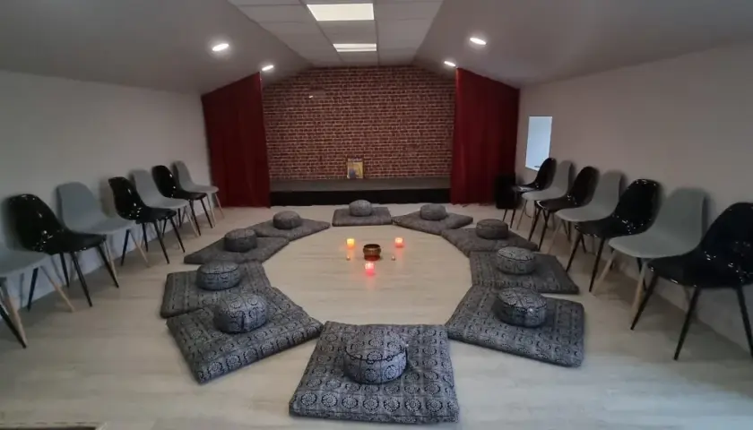 Salle de méditation avec coussins disposés en cercle et bougies allumées. Des chaises attendent autour, créant une atmosphère paisible, comme un refuge au petit goulet.