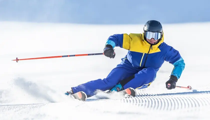 Un skieur en combinaison jaune et bleue vire sur la neige, ses skis paraboliques taillant la piste.
