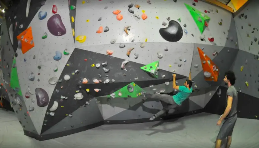 Un homme en t-shirt vert fait de l'escalade pertuis sur un mur coloré, un autre homme le regarde.