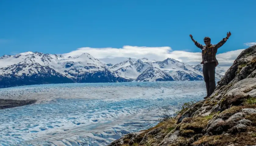 Un homme bras levés devant un glacier immense et des montagnes enneigées de la Patagonie.