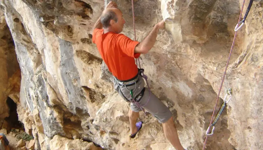 Un homme en t-shirt orange et short gris fait de l'escalade sur une paroi rocheuse. Il est assuré par une corde et semble atteindre un point élevé, un vrai **grimp azur**.