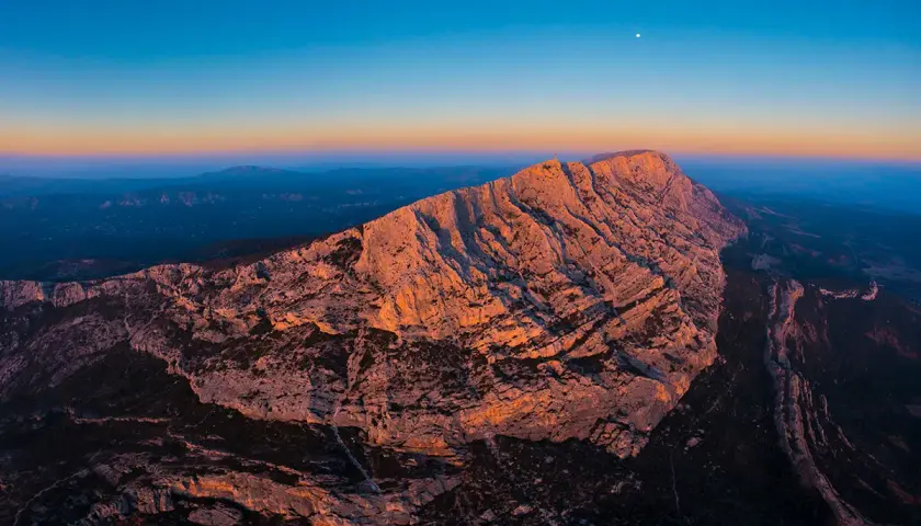 Le soleil couchant illumine la montagne Sainte-Victoire, offrant un spectacle grandiose pour les amateurs d'escalade. La lune veille sur ce paysage rocheux.
