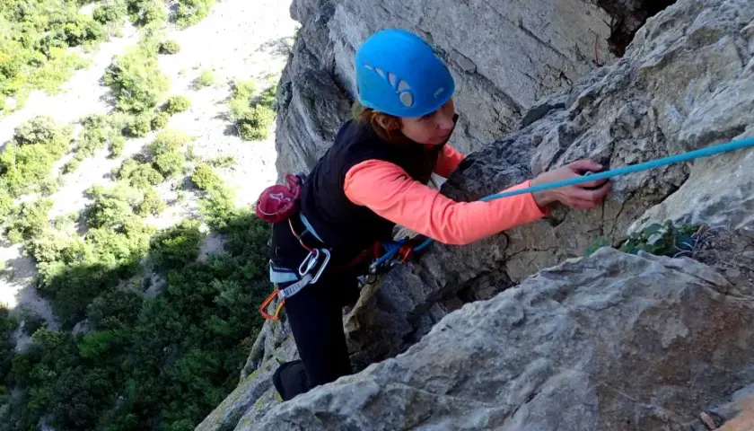 Une grimpeuse en casque bleu progresse sur une paroi rocheuse, corde bleue en main. L'escalade à Thaurac offre des vues spectaculaires.