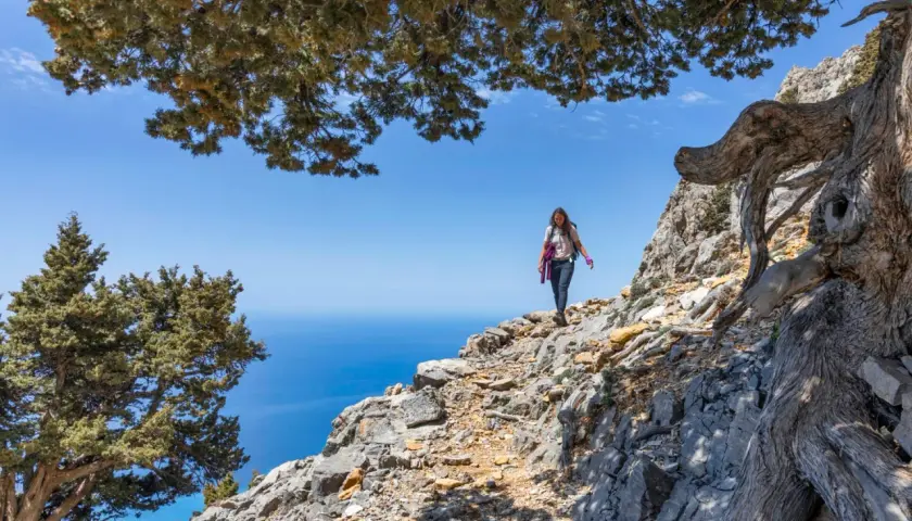 Une randonneuse explore un sentier rocailleux sur une montagne de Crète, avec la mer bleue en arrière-plan.