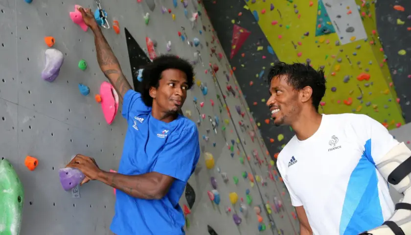 Deux grimpeurs, dont frere mawem, sourient sur un mur d'escalade coloré. L'un est en bleu, l'autre en blanc avec le logo de la France.