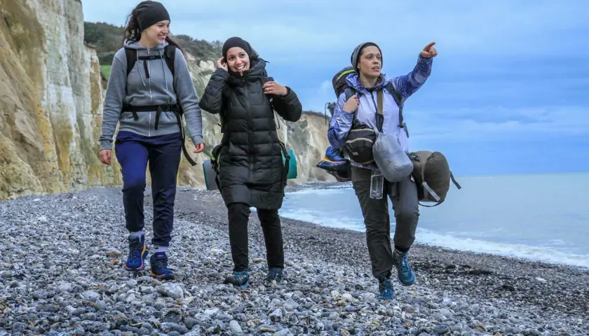 Trois randonneuses marchent sur une plage de galets, le ciel est couvert. L'une d'elles pointe le doigt vers l'horizon, peut-être pour indiquer la fin de leur GR 21 étapes.