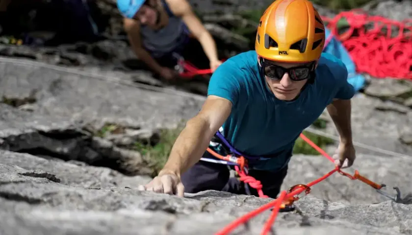 Un grimpeur concentré, casque orange, s'engage dans une voie d'escalade 64. Une corde rouge vif l'assure.