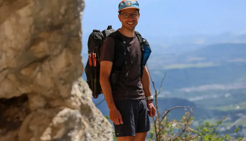 Jérémy Bigé, souriant, prêt pour l'aventure avec son sac à dos et ses bâtons de randonnée, dominant un paysage de montagnes.