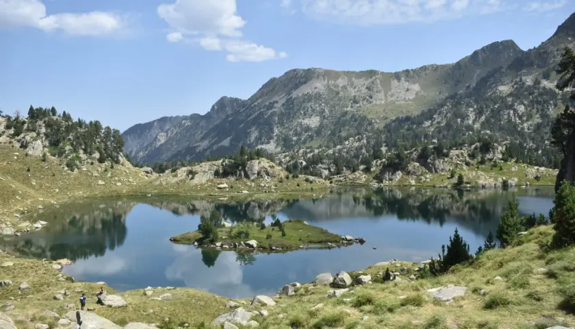 Vue paisible du cirque de Colomers, avec un lac miroir reflétant les montagnes et une petite île verdoyante.