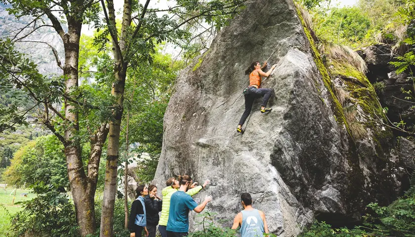 Escalade à Val di Mello : une femme grimpe un rocher pendant que des amis l'encouragent.