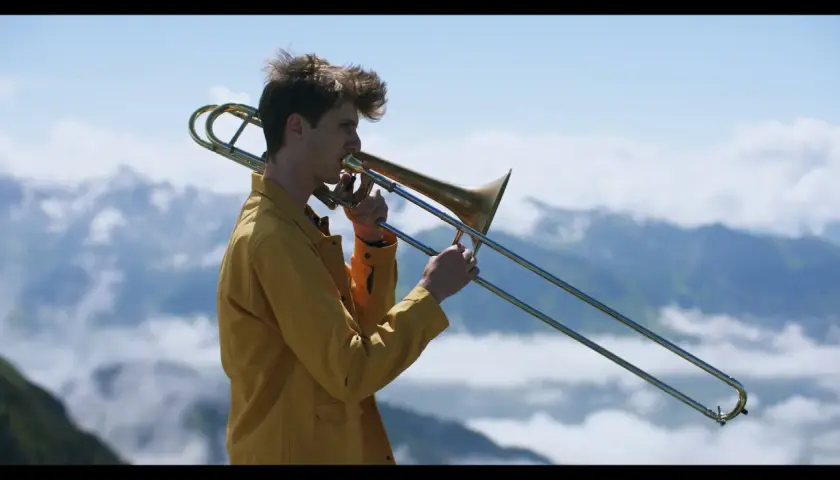 Victor Decamp Destivelle, un jeune homme aux cheveux ébouriffés, joue du trombone dans un paysage montagneux brumeux.