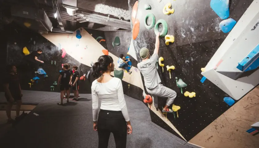 Des grimpeurs s'entraînent dans une salle de block animée, certains escaladent des murs colorés, d'autres observent.