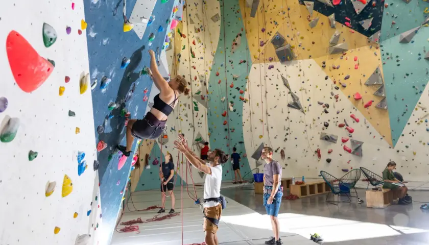 Une femme fait de l'escalade climb up sur un mur coloré, d'autres personnes observent ou se préparent.